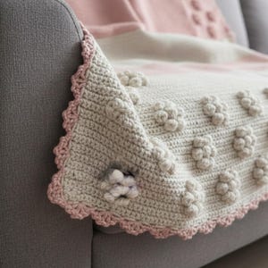 Può includere: Una coperta all'uncinetto fatta a mano con un motivo floreale. La coperta presenta una base grigio chiaro con dettagli floreali in rilievo e un bordo smerlato rosa. La coperta è drappeggiata su un divano grigio, mostrando la sua consistenza e il suo design.