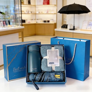 Puede incluir: Un juego de regalo azul que contiene un termo de color verde azulado, un juego de toallas dobladas y un paraguas compacto. El juego se presenta en una caja azul y una bolsa de regalo a juego. La caja tiene el texto "Best wishes".