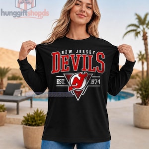 Puede incluir: Una camiseta negra de manga larga con el texto New Jersey Devils en letras rojas sobre un logotipo blanco y rojo. El gráfico incluye el texto EST. 1974 y un elemento de diseño triangular. La camiseta se presenta en un color negro sólido.