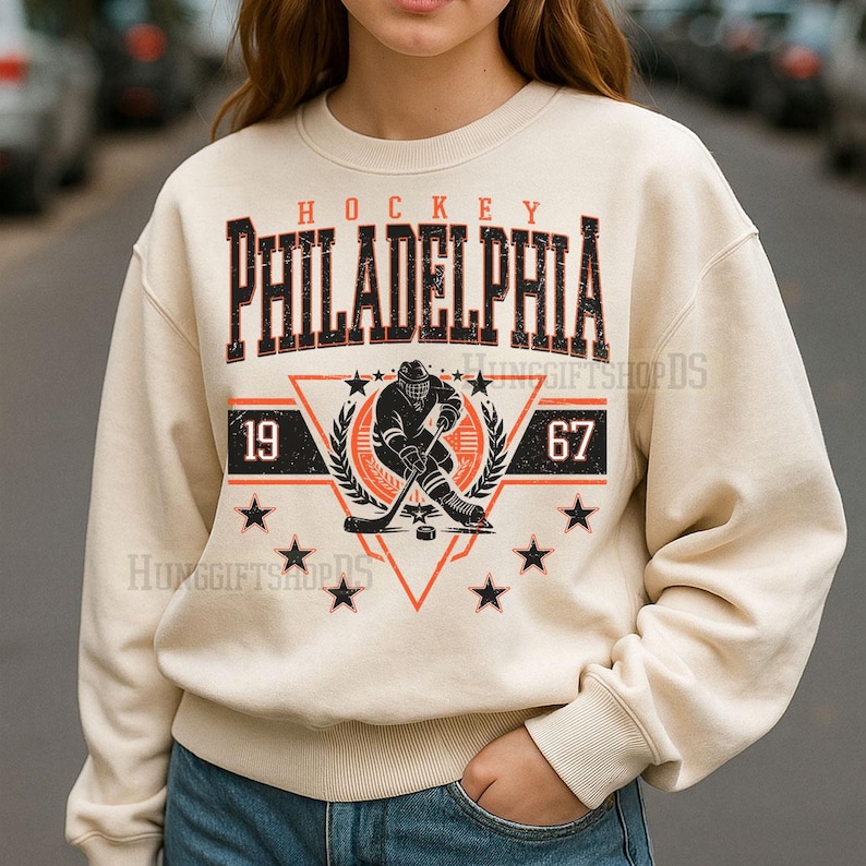 May include: Cream-colored sweatshirt with a hockey-themed graphic. The design features the word "HOCKEY" above "PHILADELPHIA" in a distressed font. Below, a hockey player illustration is centered within a triangle, flanked by the numbers "19" and "67", and surrounded by stars.