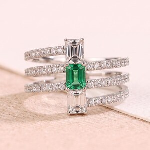 Puede incluir: Un anillo de varias bandas con tres aros plateados incrustados con pequeñas piedras claras. Una pieza central vertical conecta las bandas, mostrando una gema verde de corte esmeralda flanqueada por dos piedras claras.