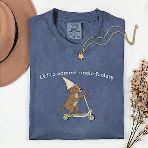 Puede incluir: Camiseta azul Comfort Colors con la frase "Off to commit uncle foolery" y un perro de dibujos animados en un scooter. También se ven un collar con colgante de estrella dorada y un sombrero marrón.