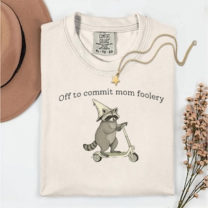 Puede incluir: Camiseta color crema con el texto "Off to commit mom foolery" y un mapache de dibujos animados montado en un scooter. El mapache lleva un sombrero de mago. También se ve un collar con colgante de estrella dorada y un sombrero marrón.