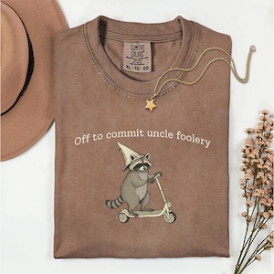 Puede incluir: Camiseta Comfort Colors marrón con el texto "Off to commit uncle foolery" y un mapache en un scooter. Un collar de estrella dorada y un sombrero marrón también son visibles.