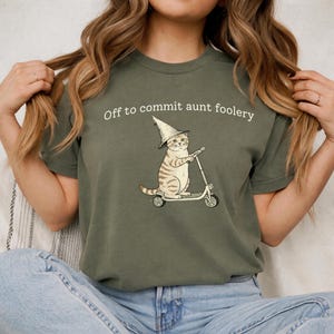 Puede incluir: Camiseta verde oliva con la frase "Off to commit aunt foolery". La camiseta presenta una ilustración caprichosa de un gato con sombrero de mago montado en un patinete. El gato es marrón y blanco con rayas negras.