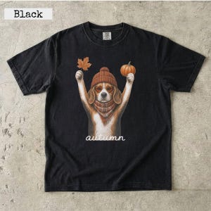 May include: A black short-sleeved t-shirt featuring a graphic of a beagle wearing a brown knit hat and plaid scarf. The dog holds a maple leaf in one paw and a pumpkin in the other. The word autumn is printed in white script below the dog.