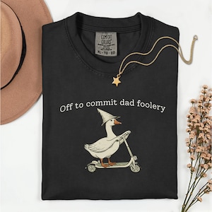 Puede incluir: Camiseta negra con la frase "Off to commit dad foolery" y un ganso de dibujos animados con un sombrero de mago montado en un patinete. También se ven un collar con colgante de estrella dorada y un sombrero marrón.