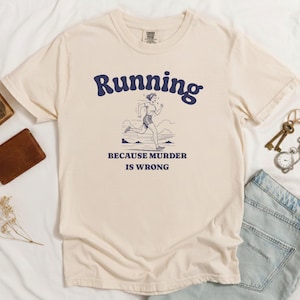 May include: A cream-colored short-sleeve t-shirt featuring a dark blue graphic of a person running. The text above the graphic reads Running and the text below reads BECAUSE MURDER IS WRONG. The shirt has a crew neckline and a standard fit.
