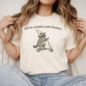 Puede incluir: Camiseta color crema con un mapache de dibujos animados montado en un patinete y con un sombrero de mago. El texto "Off to commit aunt foolery" está impreso encima del mapache. El diseño es en tonos grises y verdes.