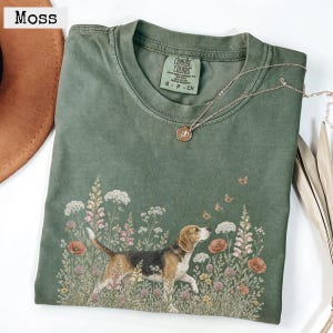Puede incluir: Una camiseta de cuello redondo color verde musgo con un estampado gráfico de un perro beagle de pie en un campo de flores silvestres y pequeñas mariposas. La prenda tiene una etiqueta Comfort Colors en el cuello y un collar de cadena dorada.