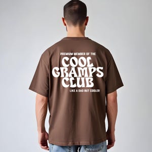 May include: A brown t-shirt with white text on the back. The text reads "PREMIUM MEMBER OF THE COOL GRAMPS CLUB LIKE A DAD BUT COOLER." The shirt is a solid colour and has short sleeves.
