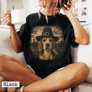 May include: A black t-shirt featuring a graphic print of a beagle dog wearing a pilgrim hat with a gold buckle. The dog has a piece of wheat in its mouth, and the background shows a field with two turkeys under a cloudy, dark sky.