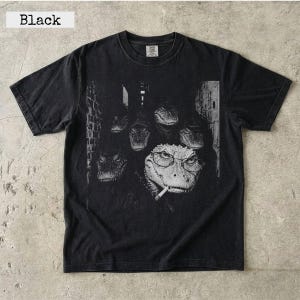 May include: A black short-sleeve t-shirt featuring a graphic print of several alligators in a dark, textured setting. One prominent alligator in the foreground wears round eyeglasses and has a cigarette in its mouth, surrounded by others.