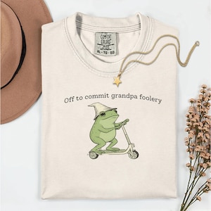 Peut inclure: T-shirt crème avec l'inscription « Off to commit grandpa foolery » et une grenouille de dessin animé sur un scooter. La grenouille porte un chapeau. Un collier doré avec un pendentif étoile et un chapeau marron sont également visibles.