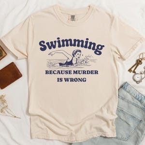 Puede incluir: Camiseta color crema con el texto azul marino "Swimming Because Murder Is Wrong". El gráfico presenta una ilustración vintage de una mujer nadando. La camiseta está hecha de un material suave y tiene un cuello redondo clásico.