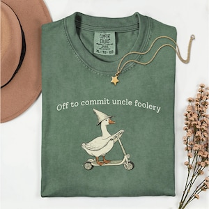 Puede incluir: Camiseta verde salvia con la frase "Off to commit uncle foolery" y un ganso de dibujos animados montado en un scooter, con un sombrero de mago. También se ven un collar con colgante de estrella dorada y un sombrero marrón.
