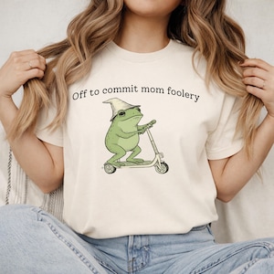 Puede incluir: Camiseta color crema con una rana verde montada en un patinete y un sombrero de mago. El texto "Off to commit mom foolery" está impreso encima de la rana. La camiseta es de material suave.