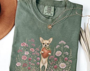 May include: A moss green crew neck t-shirt featuring a graphic of a tan chihuahua holding a large red strawberry, surrounded by pink and purple wildflowers. The shirt has a Comfort Colors label inside the collar and a thin gold chain necklace.