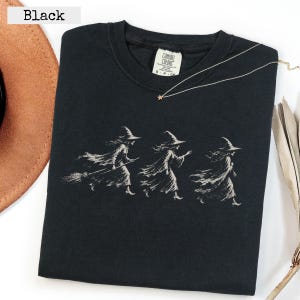 Può includere: Una t-shirt nera a maniche corte con una stampa grafica bianca raffigurante tre streghe in movimento. La prima strega porta una scopa, mentre le altre sono raffigurate con vesti fluenti e cappelli a punta.