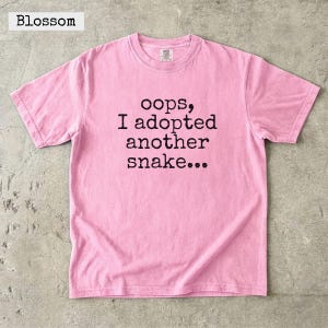 Peut inclure: Un t-shirt rose clair à manches courtes avec le texte imprimé en noir "oops, I adopted another snake..." centré sur la poitrine. Le vêtement présente une encolure ronde et une coupe standard, avec le mot "Blossom" visible en haut à gauche.