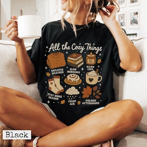 以下が含まれることがあります： All the Cozy Thingsというテキストと、セーター、本、キャンドル、シナモンロール、マグカップ、靴下、雨雲、秋の葉のイラストが描かれた黒いTシャツ。Sweater WeatherやWarm Cupsなどのラベルが添えられています。