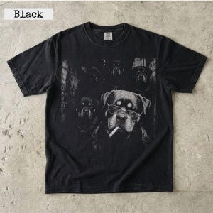 May include: A black short-sleeve t-shirt featuring a graphic print of multiple Rottweiler dogs. One prominent dog in the foreground wears wire-rimmed glasses and has a cigarette in its mouth. The design is rendered in shades of gray and white.