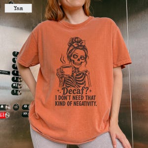 Può includere: Una t-shirt a maniche corte color arancione bruciato con una grafica nera di uno scheletro con uno chignon che tiene in mano una tazza di caffè. Il testo recita: Decaf? I DON'T NEED THAT KIND OF NEGATIVITY. La maglia ha un girocollo e una vestibilità ampia.