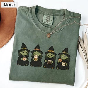 Peut inclure: Un t-shirt à manches courtes vert mousse avec un motif représentant quatre sorcières à la peau verte portant des chapeaux et des robes noirs. Chaque sorcière tient un objet lié au café, comme des tasses ou une carafe. Étiquette Comfort Colors.