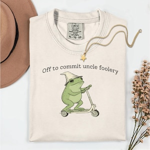 Puede incluir: Camiseta color crema con el texto "Off to commit uncle foolery". La camiseta presenta una rana de dibujos animados con sombrero montada en un patinete. También se ve un collar dorado con un colgante de estrella y un sombrero marrón.