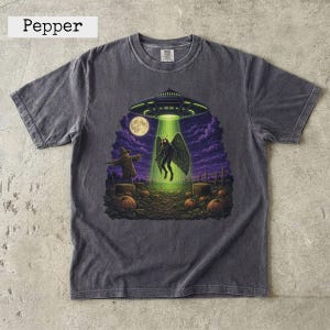 May include: A charcoal grey short-sleeve t-shirt featuring a graphic print of a moth-like creature being abducted by a UFO in a pumpkin patch at night. The scene includes a scarecrow, a full moon, and purple clouds under a bright green beam.