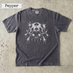 Może przedstawiać: Ciemnoszary t-shirt z krótkim rękawem z białym nadrukiem graficznym przedstawiającym biedronkę z efektem rozpryskanej farby. Koszulka ma okrągły dekolt i metkę przy kołnierzu. Słowo Pepper jest wydrukowane czarną czcionką nad koszulką.