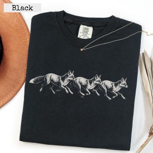 Peut inclure: Un t-shirt noir avec une impression graphique blanche représentant trois renards courant en ligne au centre. Le t-shirt a un col rond et une étiquette intérieure blanche indiquant Comfort Colors, S, et d'autres textes. Une fine chaîne dorée est posée dessus.