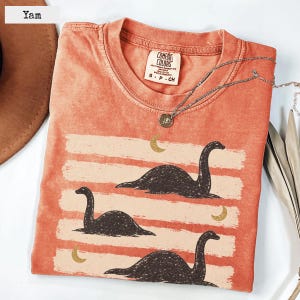 Può includere: Una t-shirt color igname con una grafica di tre creature marine scure dal collo lungo che nuotano su pennellate orizzontali color crema, accentuata da tre piccole lune crescenti dorate e un'etichetta del marchio Comfort Colors.