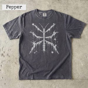 Puede incluir: Una camiseta de manga corta gris carbón con un estampado gráfico blanco de un insecto palo. El diseño tiene un efecto de pintura salpicada y goteante. La camiseta tiene un cuello redondo y una etiqueta con el texto Pepper.