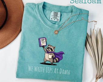 May include: A seafoam green t-shirt featuring a graphic of a raccoon wearing a purple cape, holding a coffee mug and a purple binder labeled IEP Individualized Education Program. The text below reads We Write IEPs At Dawn in white lettering.