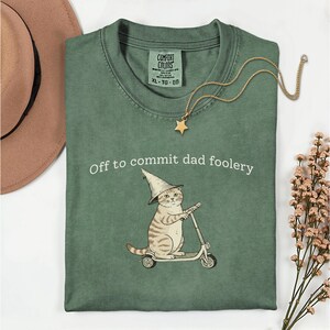 Puede incluir: Una camiseta verde con la frase "Off to commit dad foolery" y un gato de dibujos animados en un patinete, con un sombrero de mago. También se ve un collar dorado con un colgante de estrella y un sombrero marrón.