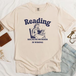 May include: Cream-colored t-shirt with a navy blue graphic. The graphic features the word "Reading" above an illustration of a woman reading a book, with the text "BECAUSE MURDER IS WRONG" below. The shirt is folded.