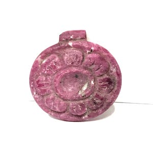 May include: A circular, deep pink gemstone pendant carved with a central flower motif. The stone features a textured, matte surface with natural inclusions and a small, integrated loop at the top for suspension, showcasing a vibrant magenta hue.