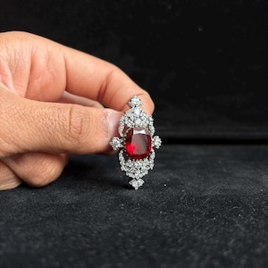 May include: A brooch featuring a large, central, cushion-cut deep red gemstone surrounded by a decorative silver-toned metal frame encrusted with numerous small, clear, sparkling white stones arranged in an intricate, symmetrical filigree pattern.