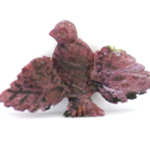 May include: A carved bird figurine with outstretched wings. The bird is a deep maroon color with hints of green and black. The wings are detailed with feather-like patterns. The figurine is set against a white background.