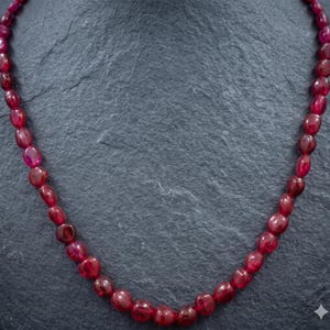 Può includere: Una collana composta da numerose pietre preziose ovali, di colore rosso intenso. Le pietre sono infilate insieme, formando un anello continuo. La collana è esposta su uno sfondo scuro e strutturato, che mette in risalto il colore ricco e la lucentezza delle pietre.