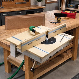 Könnte beinhalten: Ein Frästisch aus hellem Holz mit einer flachen Oberfläche, integrierten Metallschienen, einem zentralen schwarzen runden Einsatz, einem verstellbaren Anschlagsystem, einem grünen Absaugschlauch und einem T-Griff-Schlüssel auf der Tischplatte.