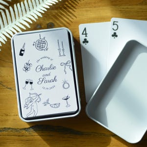 May include: A white rectangular metal tin containing a deck of playing cards with a custom design. The lid features black line art including a disco ball, a ring, a cupid, and text reading The wedding of Charlie and Sarah 12.19.2026.