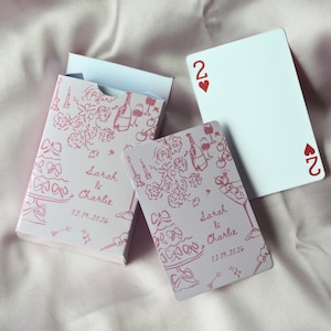 Può includere: Un mazzo di carte da gioco e la sua scatola, con un design rosa chiaro con illustrazioni di fiori, torta e champagne. Le carte e la scatola sono personalizzate con i nomi "Sarah & Charlie" e la data "12.19.2026". Una carta mostra il numero "2" e un cuore.