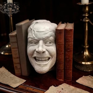 May include: A white, sculpted face with a wide, unsettling grin, positioned between several antique books. The books have aged, reddish-brown covers. The image has a dark, gothic aesthetic, with a logo in the upper left corner.