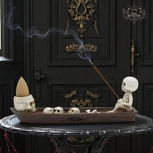 Puede incluir: Un portaincienso decorativo con una figura de esqueleto sentado en un bote de madera, sosteniendo una varilla de incienso. El bote contiene varias calaveras pequeñas y un soporte para cono de incienso con forma de calavera. El humo se eleva del incienso. El texto "Dark Reliquary" es visible.