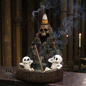May include: A decorative incense burner shaped like a mountain with a skull on top. Two small skeleton figures sit in a bowl at the base. Smoke rises from the incense, creating a mystical effect. The burner is dark brown and green.