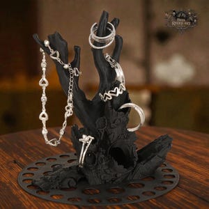 Pode incluir: Um suporte de joias preto em forma de mão com vários anéis de prata e uma pulseira. O suporte tem um design orgânico texturizado e uma base circular. O logotipo Dark Reliquary é visível no fundo.