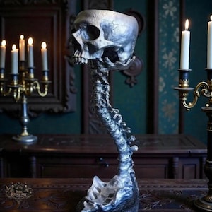 Puede incluir: Una escultura de calavera plateada con un cuello en forma de columna vertebral. La calavera tiene cuencas de los ojos detalladas, una cavidad nasal y dientes. La escultura se exhibe sobre una superficie de madera oscura, con velas encendidas al fondo. De Dark Reliquary.
