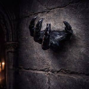 Puede incluir: Una mano negra con garras emerge de una pared de piedra gris. La mano tiene garras largas y afiladas y una superficie texturizada, creando una estética gótica y espeluznante. La imagen es oscura y dramática, ideal para la decoración de Halloween.